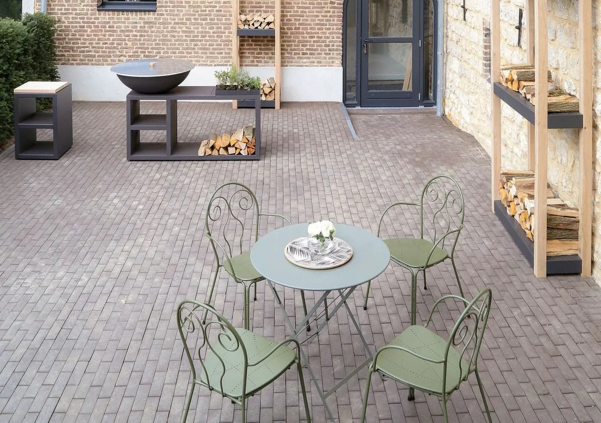 Cour pavée avec un brasero Quoco sur meuble noir au fond et une table de jardin verte au premier plan.