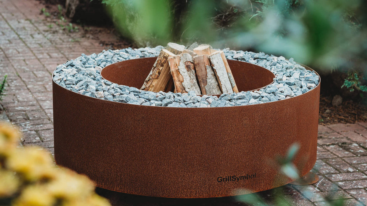 Brasero rond en acier Corten chargé de bûches, installé dans un jardin avec des feuilles jaunes au premier plan.