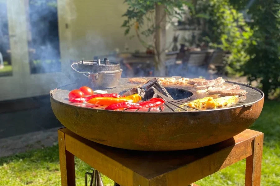 Légumes et grillades sur la plaque d’un brasero BAKKA en fonctionnement dans un jardin.