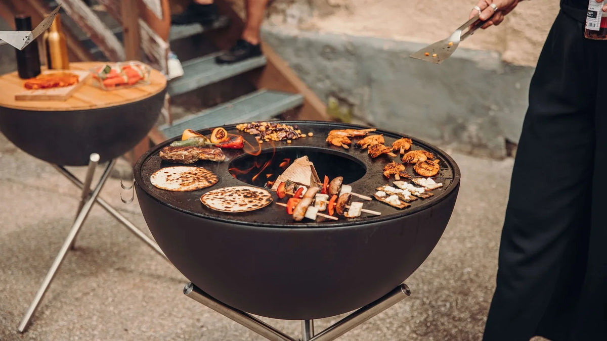Aliments répartis sur la plancha en anneau d’un BOWL 70, avec l’ouverture centrale et le bois du foyer visibles.