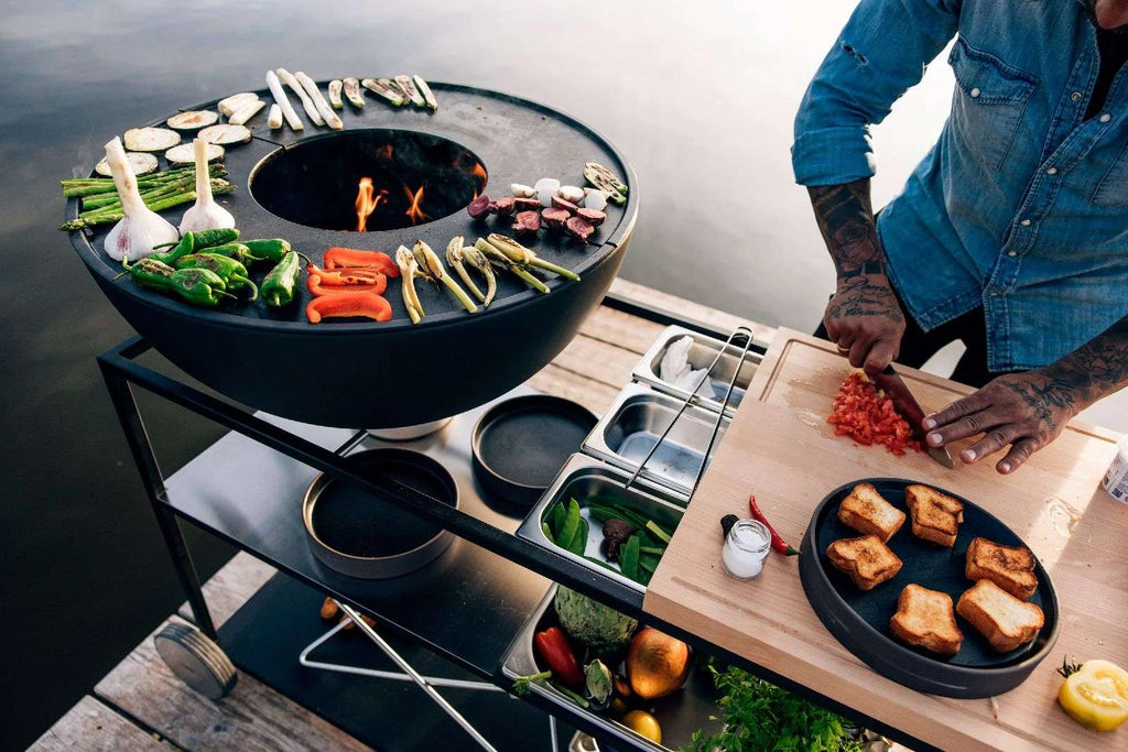 Gril circulaire noir avec une flamme et des légumes dessus, conçu pour la cuisson en extérieur.