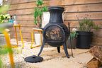Chiminea en fer forgé noir avec un devant en maille et des pieds trépied, dotée d’un corps arrondi et d’un haut évasé.