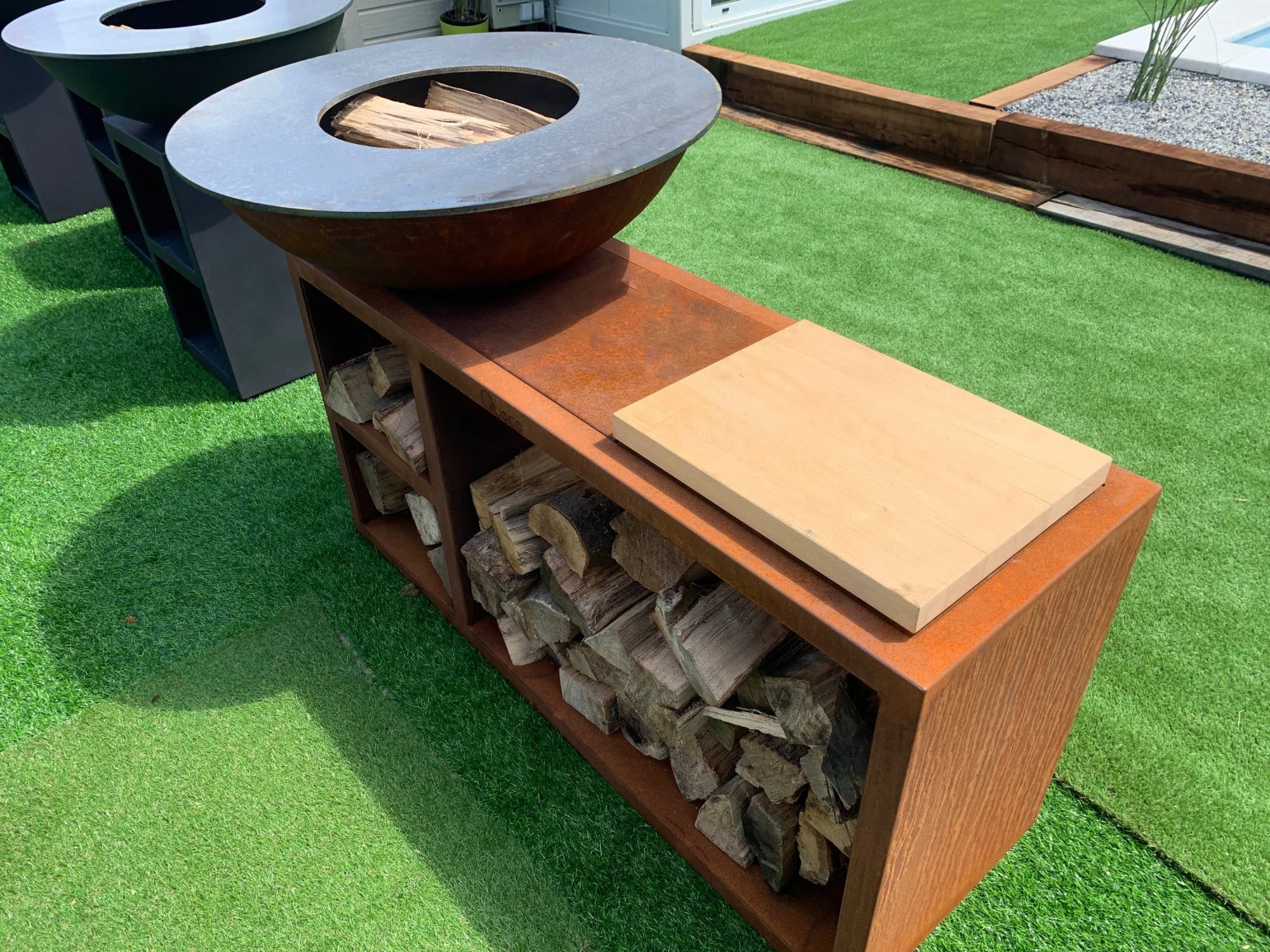 Bol en cuve de foyer de ton cuivré rustique avec un dessus circulaire gris foncé, reposant sur une unité d’étagères ouverte remplie de bûches empilées.