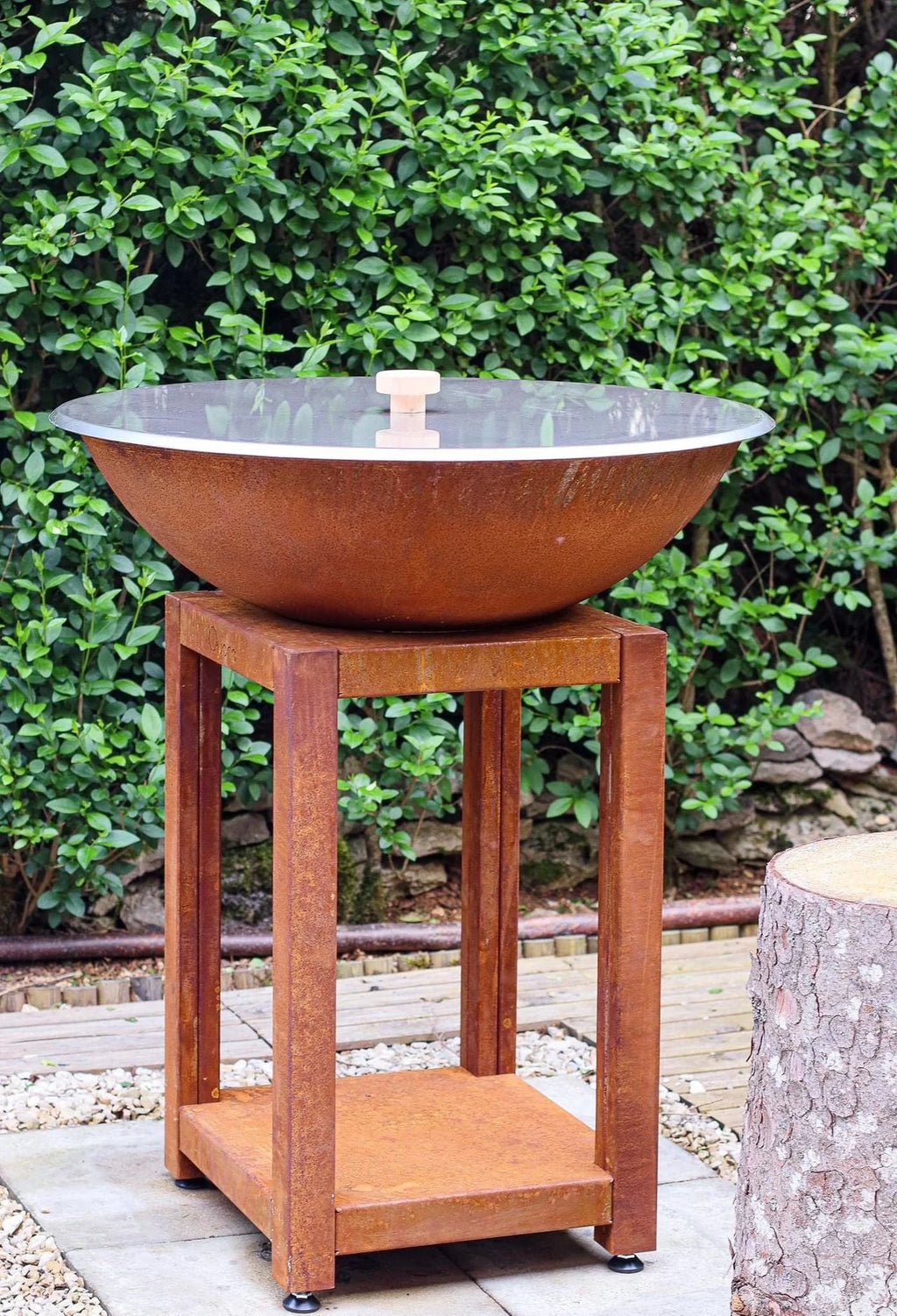 Bol d’acier rouillé de couleur cuivre patiné avec un couvercle en verre transparent et une poignée en bois, reposant sur un support en métal rouillé assorti.