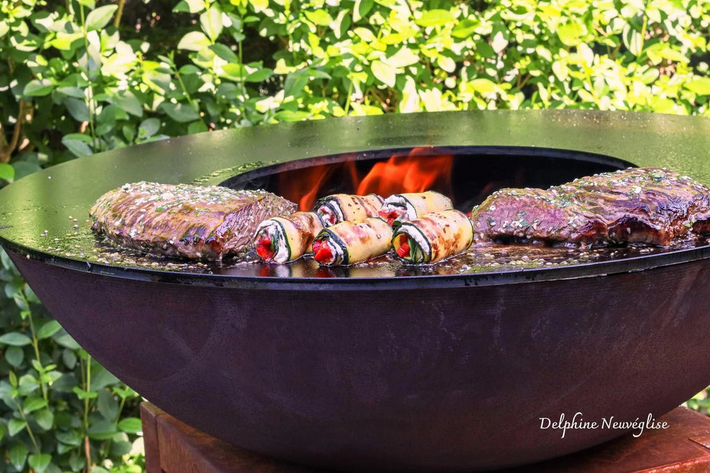 Foyer d’extérieur en fonte noire avec des steaks saisies et des pains farcis en train de griller sur des flammes ouvertes.