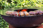 Foyer d’extérieur en fonte noire avec des steaks saisies et des pains farcis en train de griller sur des flammes ouvertes.