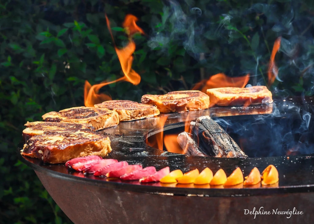 Tranches de pain perdu dorées-brunes qui grésillent sur des flammes nues sur une grille en fonte noire, accompagnées de lanières de bacon rose disposées avec soin et de fruits citriques jaunes en tranches.