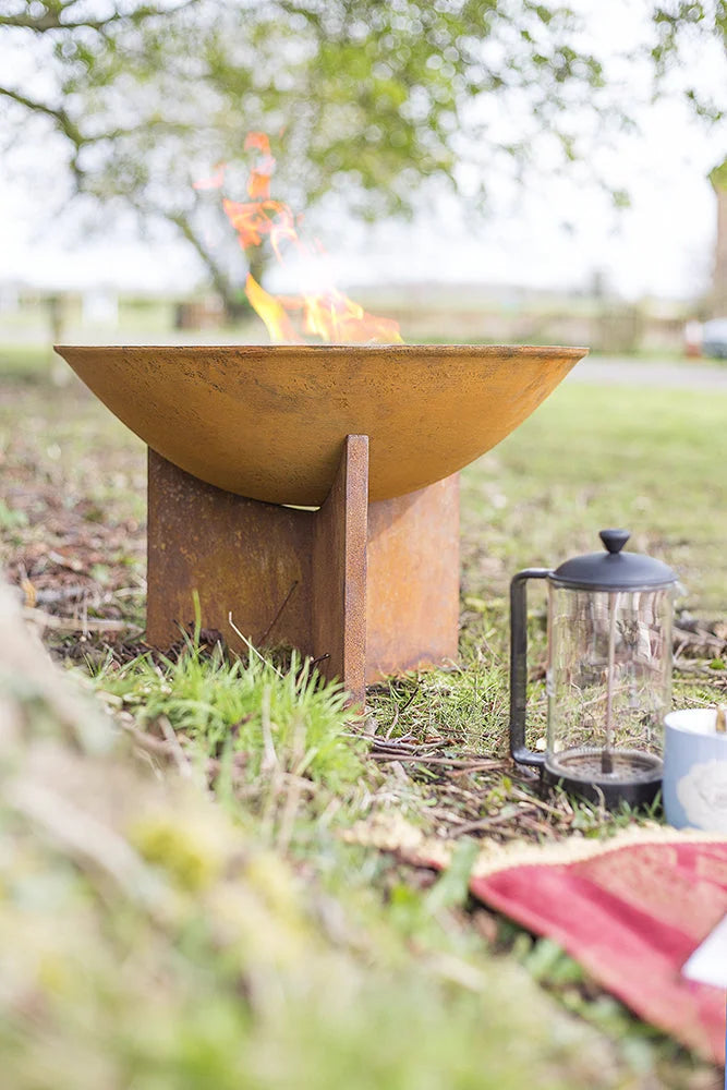 Bol en acier rouillé avec une flamme brûlant à l’intérieur, reposant sur une surface herbeuse en extérieur.