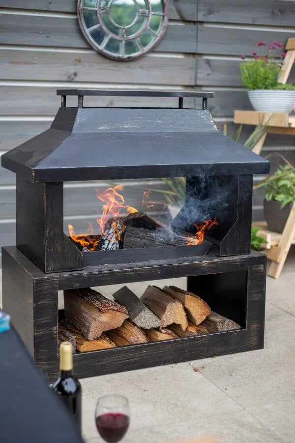 Cheminée extérieure en fer forgé noir avec un front en verre, abritant des bûches en combustion et du bois empilé en dessous.