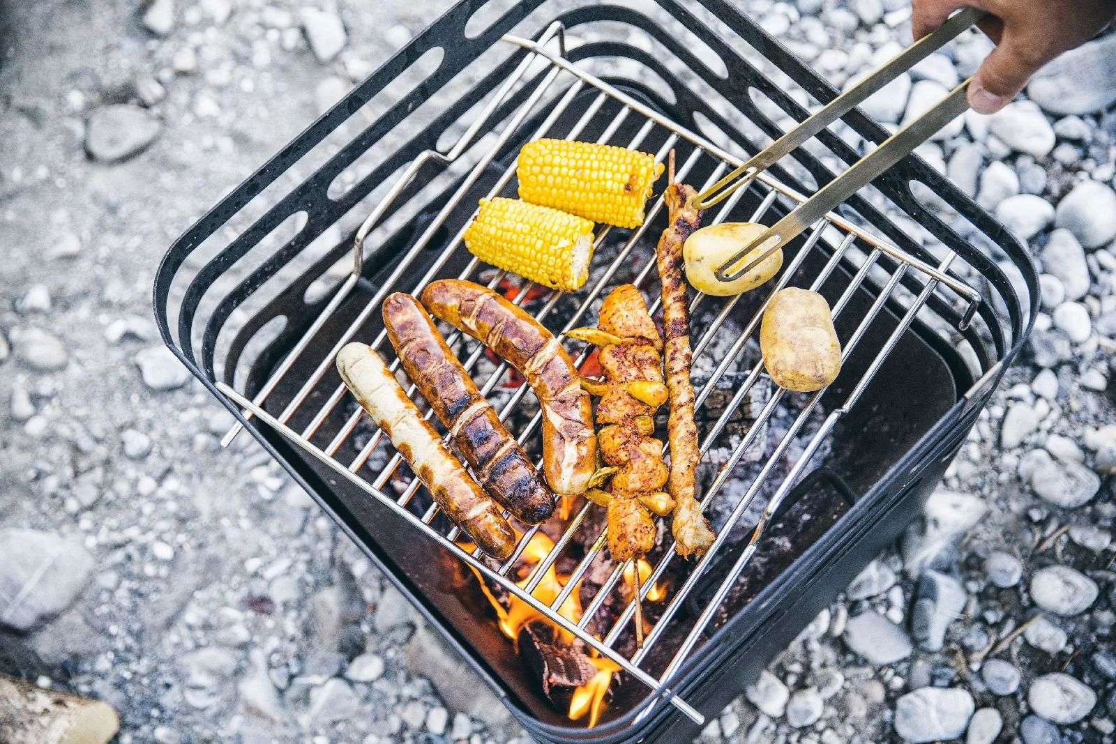 Barbecue au charbon avec grille en métal noir et poignée, cuisant des saucisses, du maïs en épi, des pommes de terre et des brochettes au-dessus de charbons incandescents.