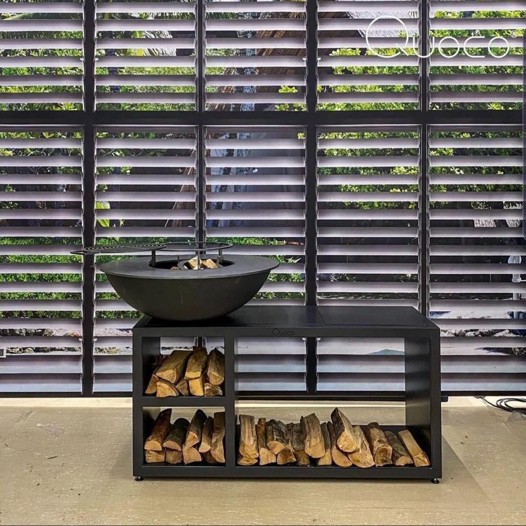 Bol en fonte noire pour foyer avec grille de barbecue intégrée, reposant sur un support en métal noir assorti qui maintient des bûches empilées en dessous.