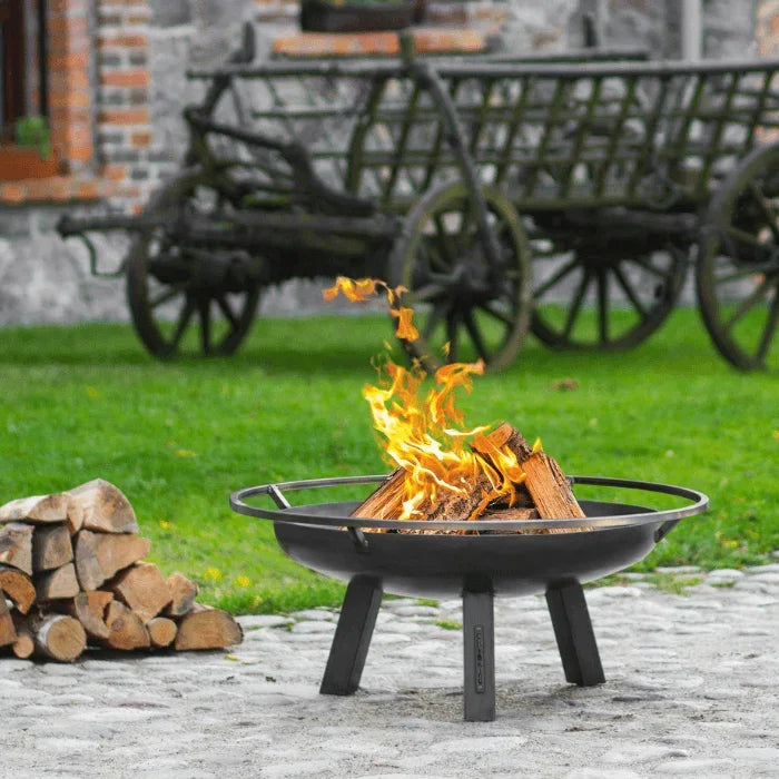 Foyer de feu en métal noir avec trois pieds et un design circulaire, présentant des bûches en train de brûler à l’intérieur.