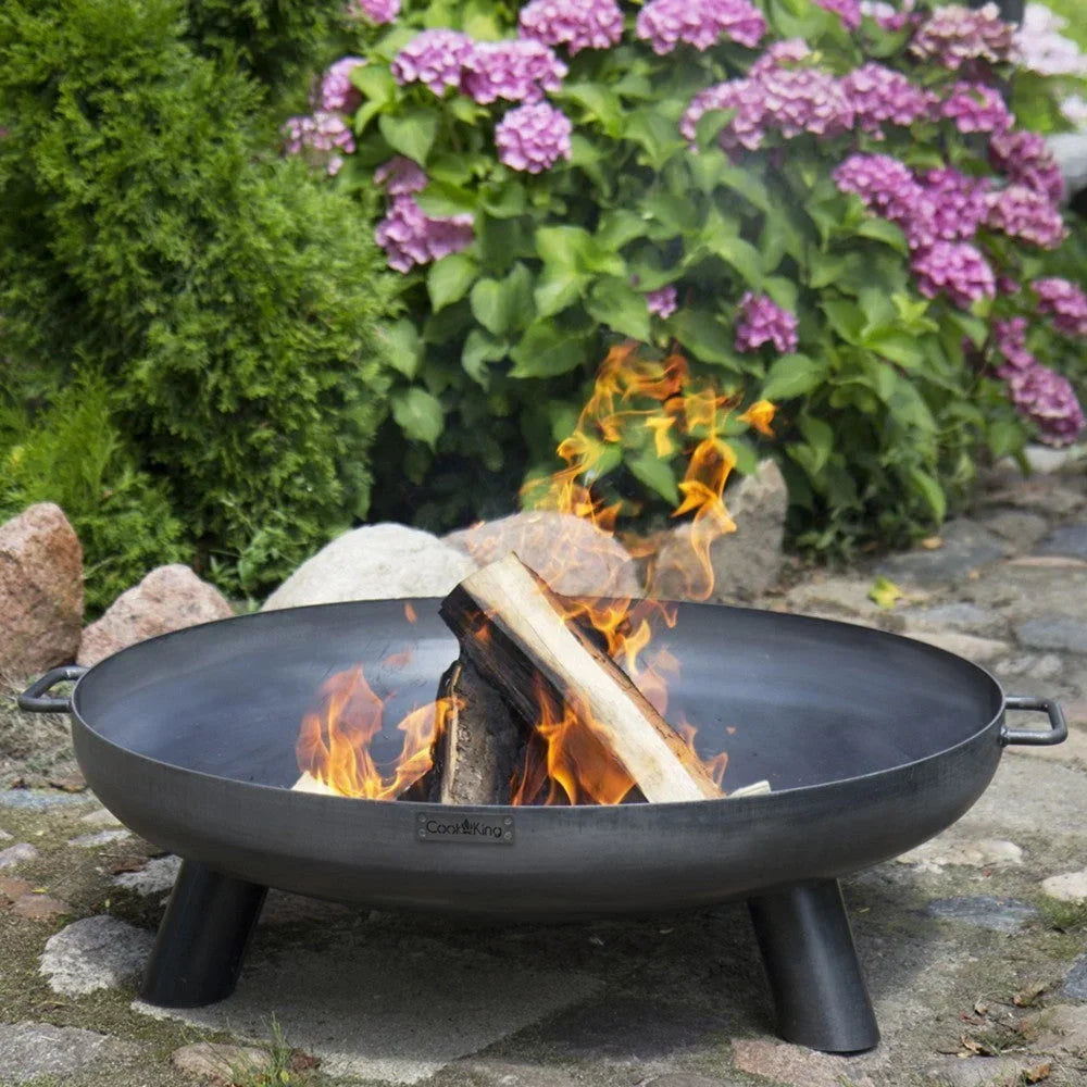 Fosse à feu noire avec des bûches de bois en train de brûler à l’intérieur, entourée de fleurs violettes et de feuillage vert.