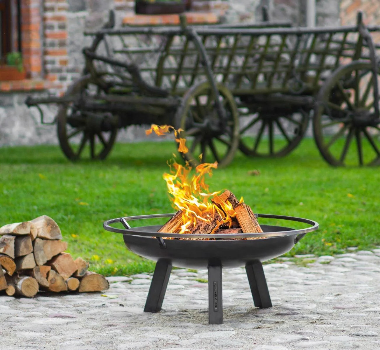 Foyer de feu en métal noir avec trois pieds et un design circulaire, présentant des bûches qui brûlent à l’intérieur.