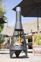 Foyer de feu en métal noir avec une cheminée en argent et un écran en maille, présentant un design conique sur une terrasse sous un parasol