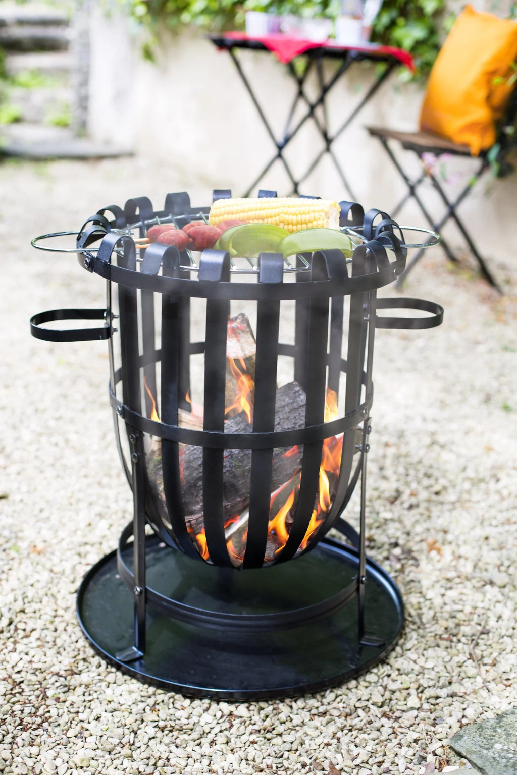 Foyer de feu en fer forgé noir avec un gril circulaire en haut retenant du maïs et des légumes au-dessus de braises rougeoyantes.