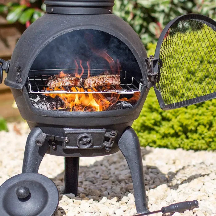 Chimenea en fonte noire avec une porte en maille ouverte pour révéler des braises incandescentes et des côtelettes en train de cuire sur une grille en fil de fer.