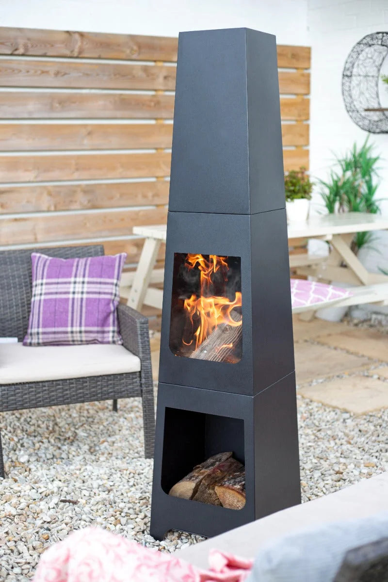 Cheminée extérieure en métal noir avec un design empilé haut, doté de panneaux de verre et de flammes visibles.