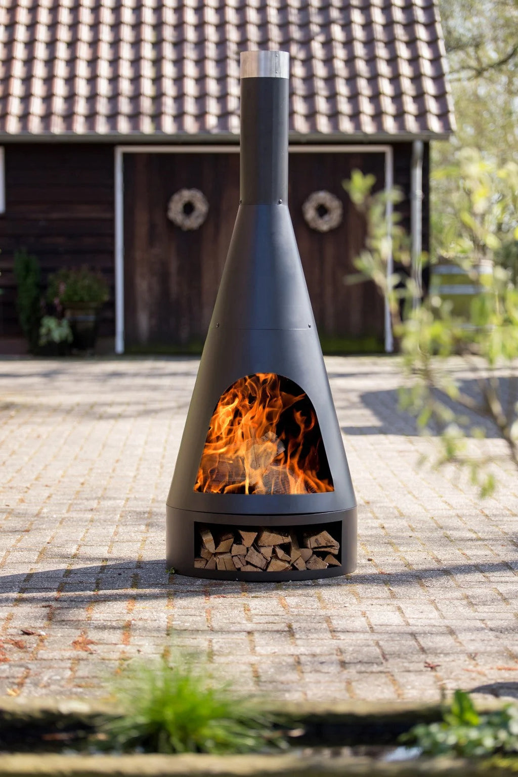 Cheminée extérieure conique en métal noir avec flammes visibles et bois empilé en dessous