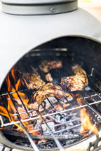 Des côtes grillées crépitent au-dessus de charbons incandescents à l’intérieur d’un gril à cheminée en céramique grise.