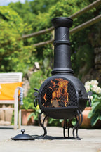 Chimenea en fer forgé noir avec une cheminée haute et une flamme visible à travers son devant en maille, reposant sur quatre pieds courbés.