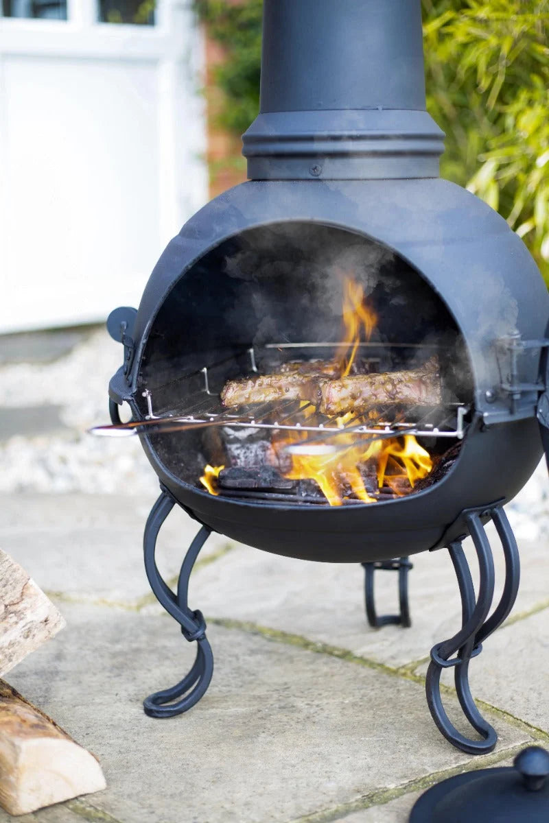 Chimenea en fer forgé noir avec un corps rond, trois pattes courbées et un feu ouvert brûlant à l’intérieur.
