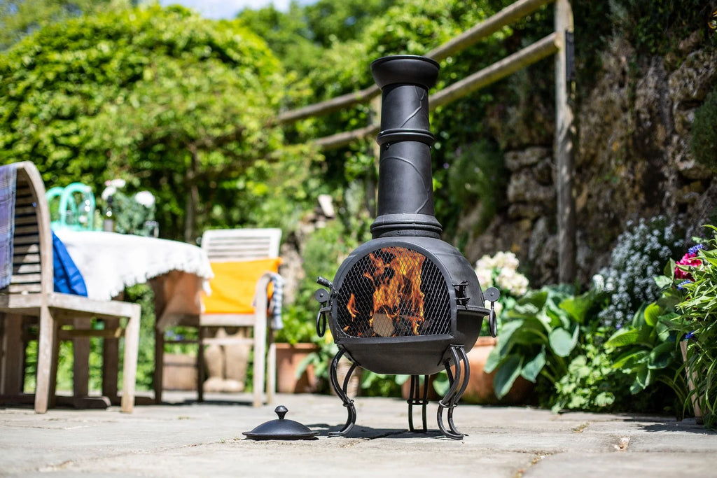Chimenea en fer forgé noir avec une flamme à l’intérieur, reposant sur trois pieds et dotée d’une cheminée haute.