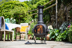 Chimenea en fer forgé noir avec une flamme à l’intérieur, reposant sur trois pieds et dotée d’une cheminée haute.