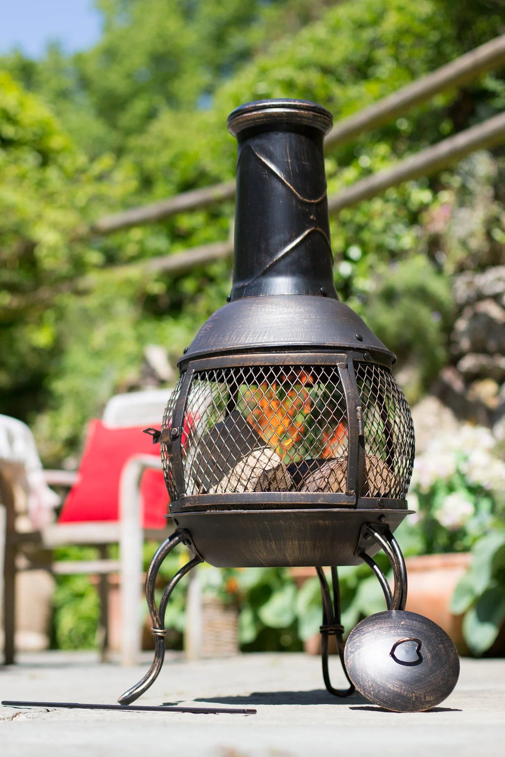 Chimenea en fonte noire avec un écran en maille et des motifs décoratifs torsadés sur sa cheminée, dotée de trois pieds courbés et d’un couvercle assorti posé à côté.