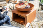Bol en acier rouillé avec une base carrée assortie, présentant un design ouvert pour les bûches et les flammes.