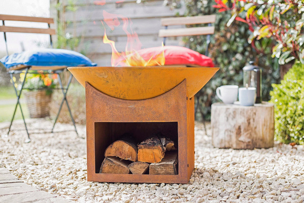Foyer de feu en acier orange rouillé avec un dessus courbé et des bûches empilées à l’intérieur