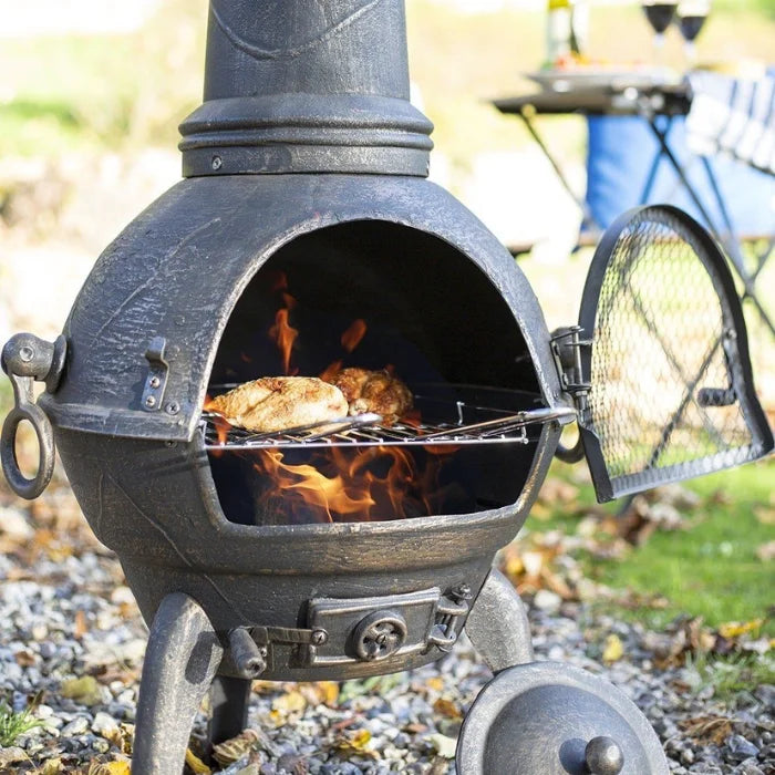 Chimenea en fonte noire avec un corps rond, porte ouverte révélant des flammes et du poulet grillé à l’intérieur.