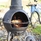 Chimenea en fonte noire avec un corps rond, porte ouverte révélant des flammes et du poulet grillé à l’intérieur.