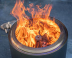 Un foyer de feu cylindrique en métal gris foncé avec une flamme orange luisante qui danse à l’intérieur et un brûleur central.