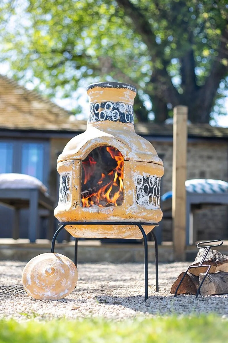 Une cheminée en céramique jaune avec des motifs circulaires noirs et un feu visible à l’intérieur, posée sur un support métallique en extérieur.
