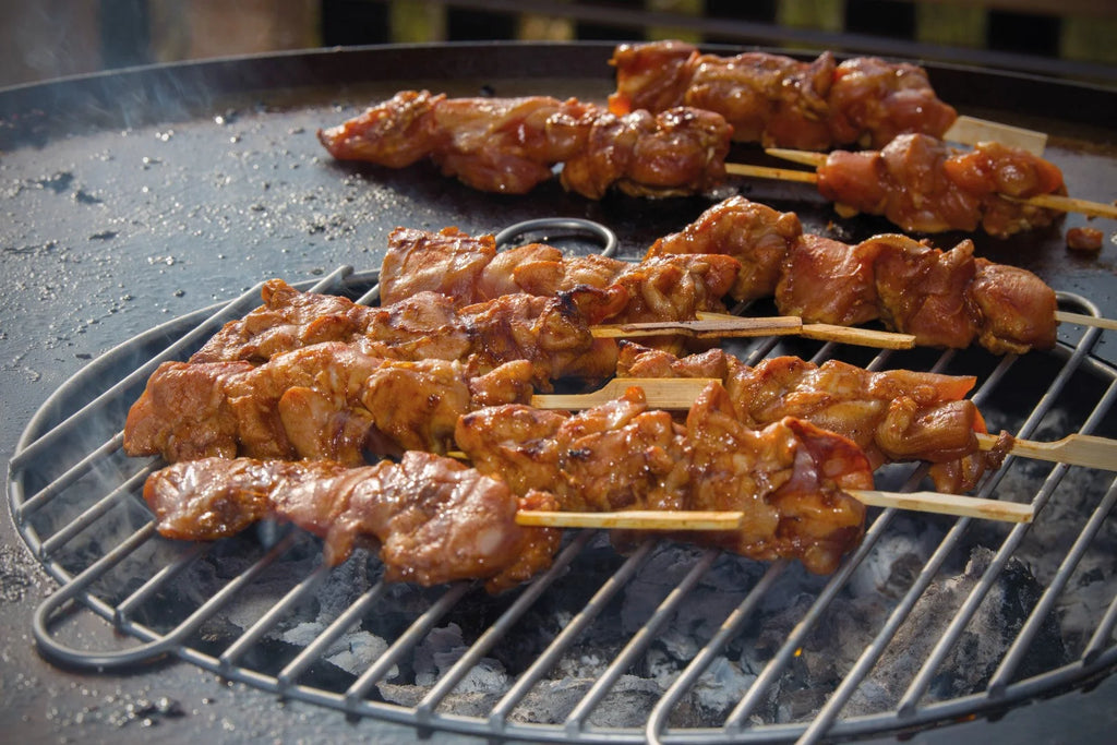 Brochettes de poulet grillé avec un glaçage brillant, brun foncé, qui grésille sur une grille en métal au-dessus de charbons incandescents.