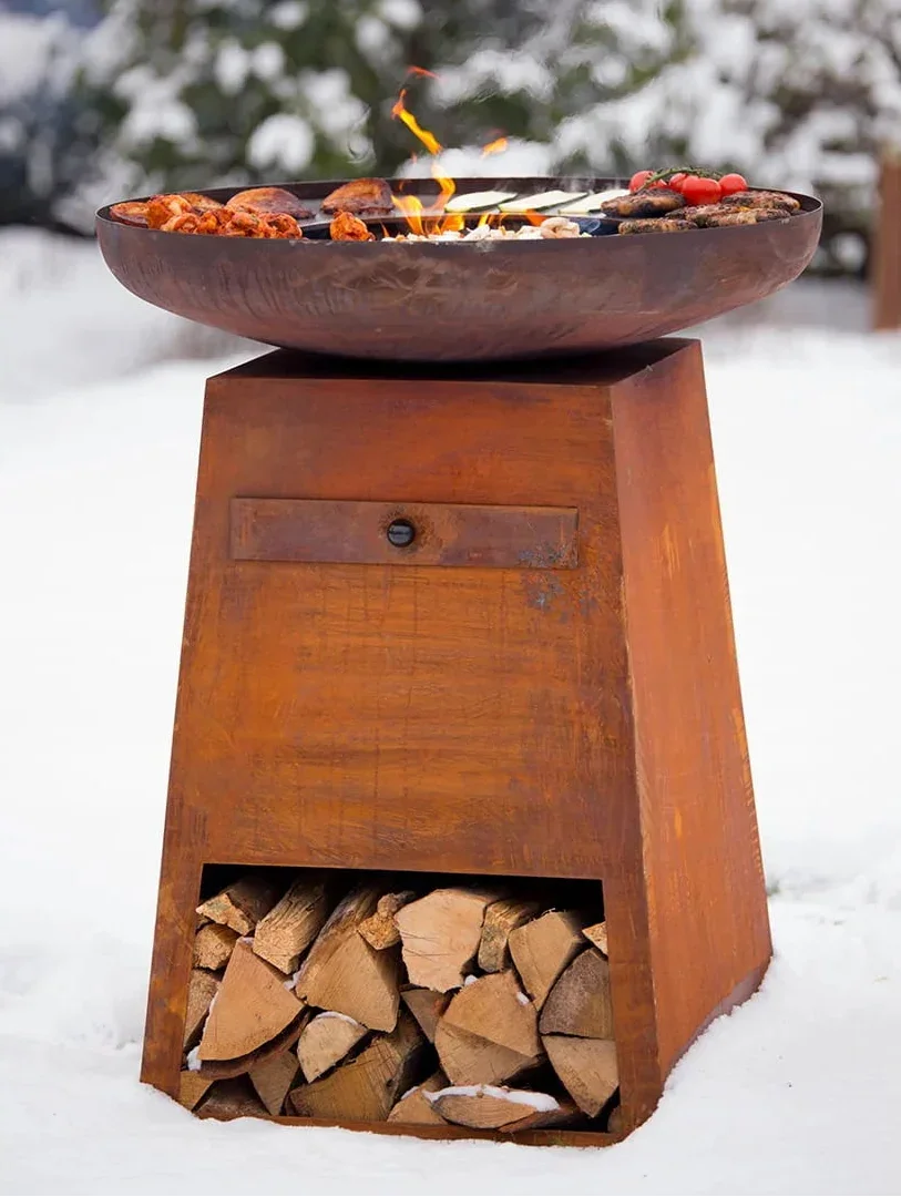 Foyer de feu en acier rouillé de couleur cuivre avec une surface de cuisson en forme de bol et un compartiment de rangement pour bois empilé en dessous