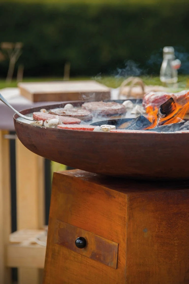 Gril rond de couleur cuivre rustique avec des flammes et de la fumée s’élevant du charbon en combustion, cuisant des galettes de viande et des champignons