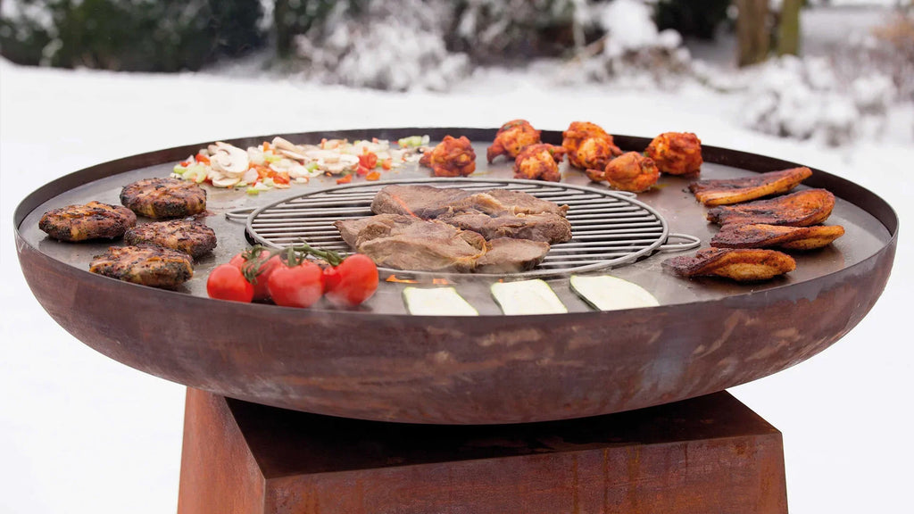 Gril rond en acier rustique avec une grille centrale en fil et divers viandes, légumes et fromages disposés autour de son bord.