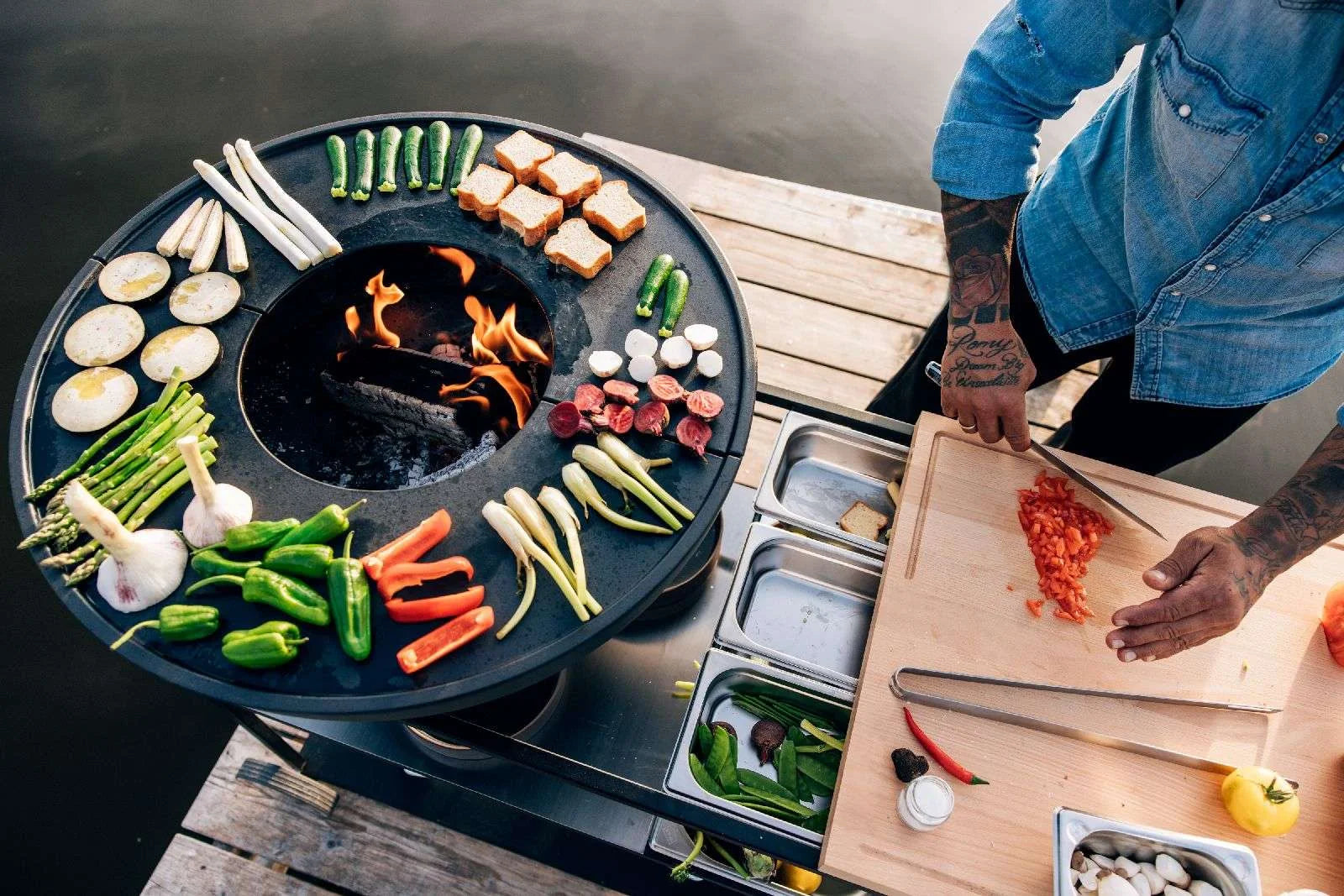 Gril rond noir avec des légumes colorés et de la viande disposés autour, présentant un design de flamme central.