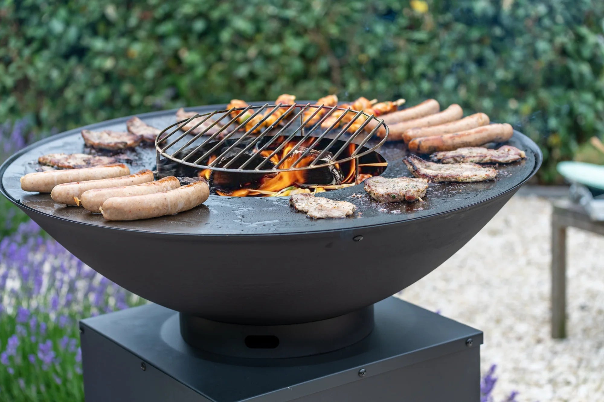 Gril rond noir avec un feu central et une grille en métal, cuisant des saucisses et des galettes de viande