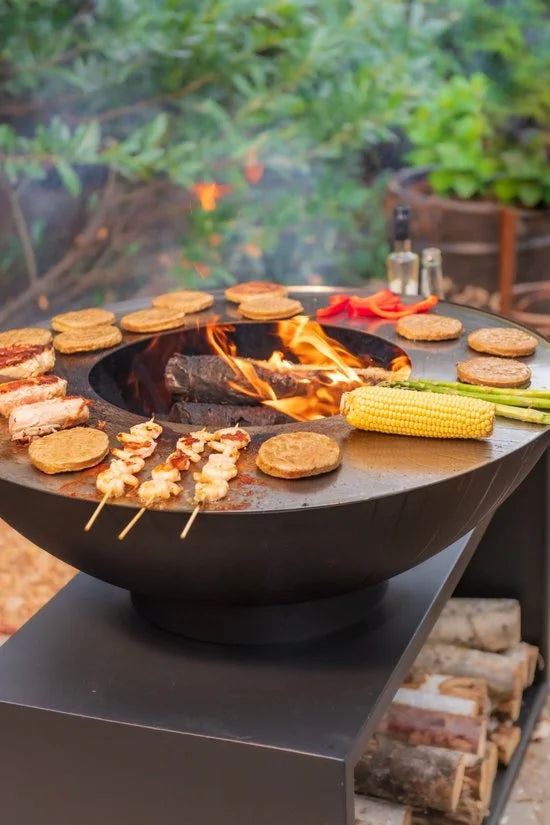 Foyer de feu en acier noir avec une surface de cuisson circulaire et une base de rangement pour bois intégrée