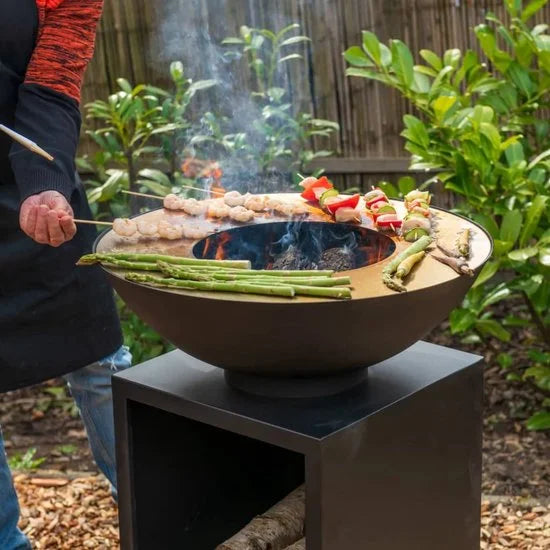 Bol en bronze rustique pour foyer avec une base noire élégante, présentant une flamme ouverte et de la fumée s’élevant du centre pendant qu’il grille des viandes et des légumes en brochettes.