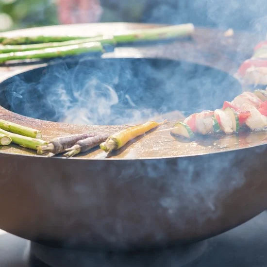 De la fumée s’élève d’un foyer de feu rond en bronze sombre tandis qu’il grille des légumes colorés et de la viande en brochettes.