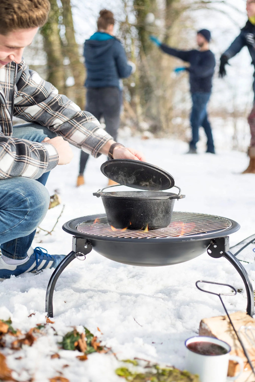 Marmite en fonte noire avec un couvercle assorti, reposant sur un gril portable au-dessus d’un feu ouvert dans un cadre extérieur enneigé