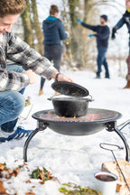 Marmite en fonte noire avec un couvercle assorti, reposant sur un gril portable au-dessus d’un feu ouvert dans un cadre extérieur enneigé