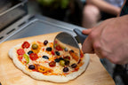 Un coupe-pizza en acier inoxydable avec une poignée ergonomique noire tranche une pizza fraîchement cuite, garnie de légumes colorés.