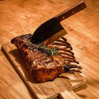 Planche à découper en bois avec un carré d’agneau et un rameau de romarin, couteau à manche brun foncé reposant dessus.