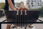 Grille mate noire avec trois brochettes attachées, probablement faite en acier inoxydable et manches en bois.