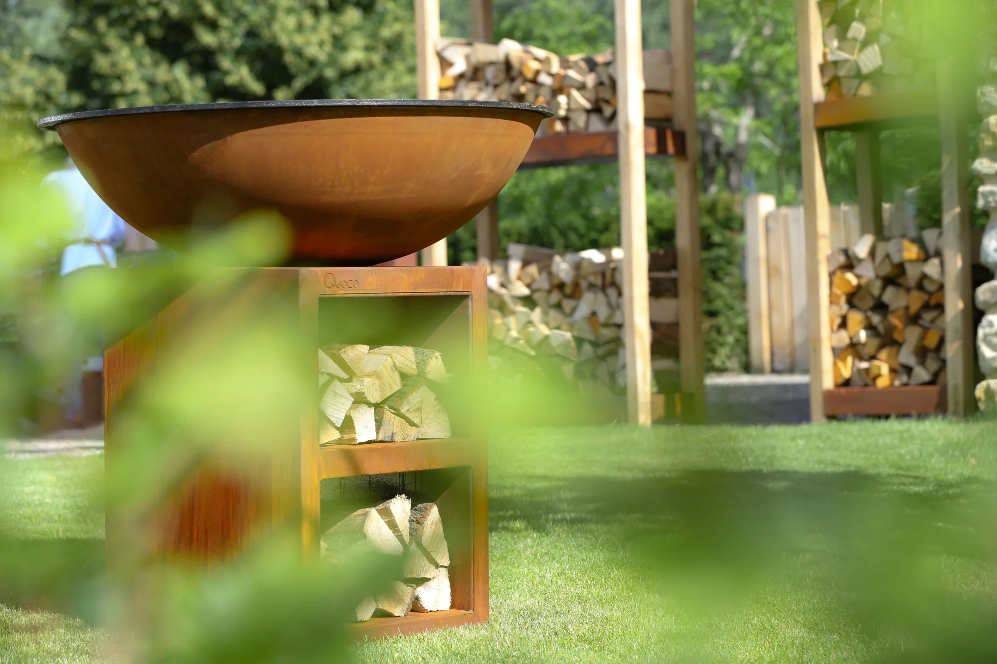 Bol en acier rouillé pour feu avec un support en bois tenant des bûches empilées.